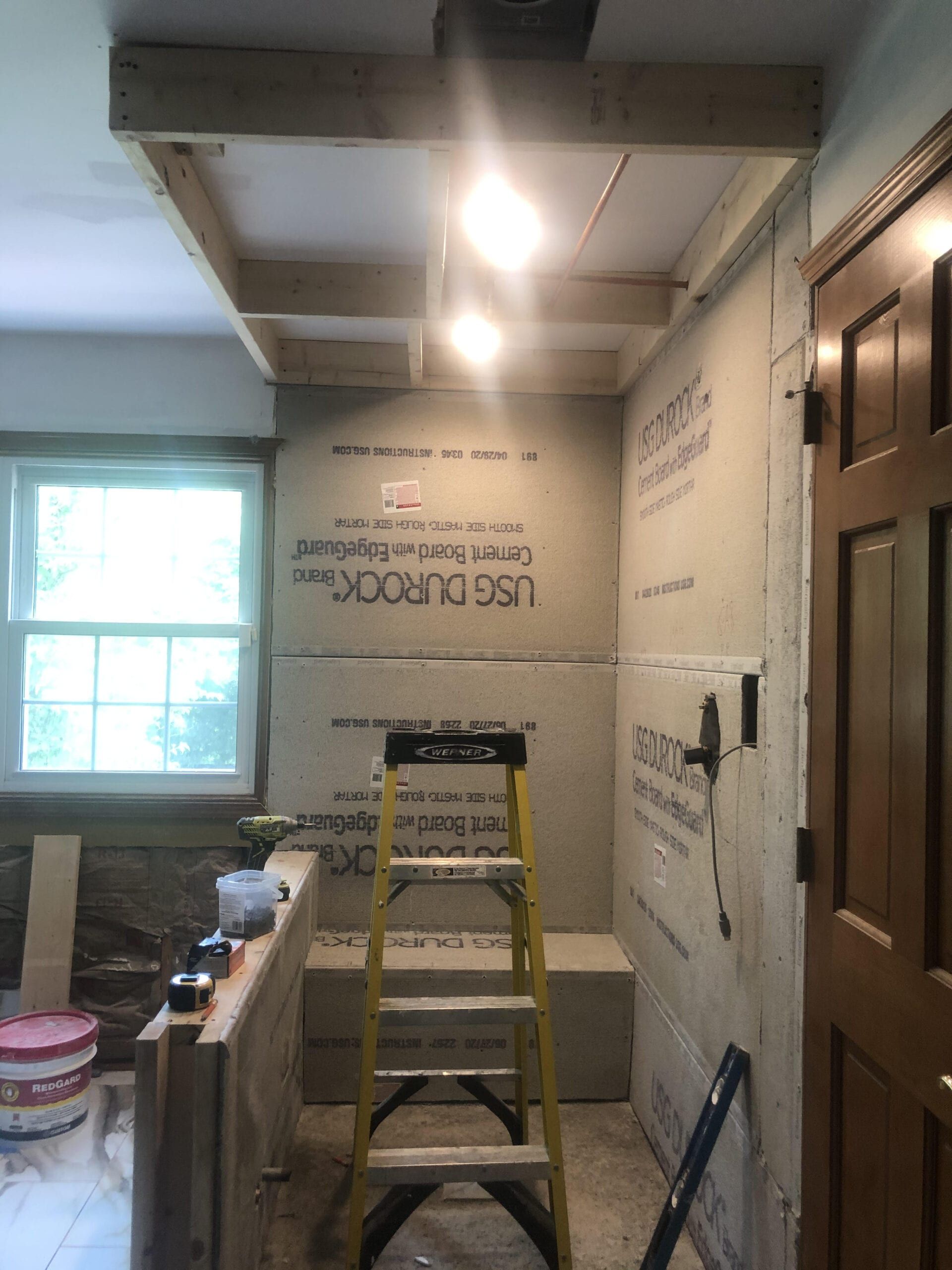 Bathroom under renovation with ladder, exposed insulation, and a window by a wooden door.