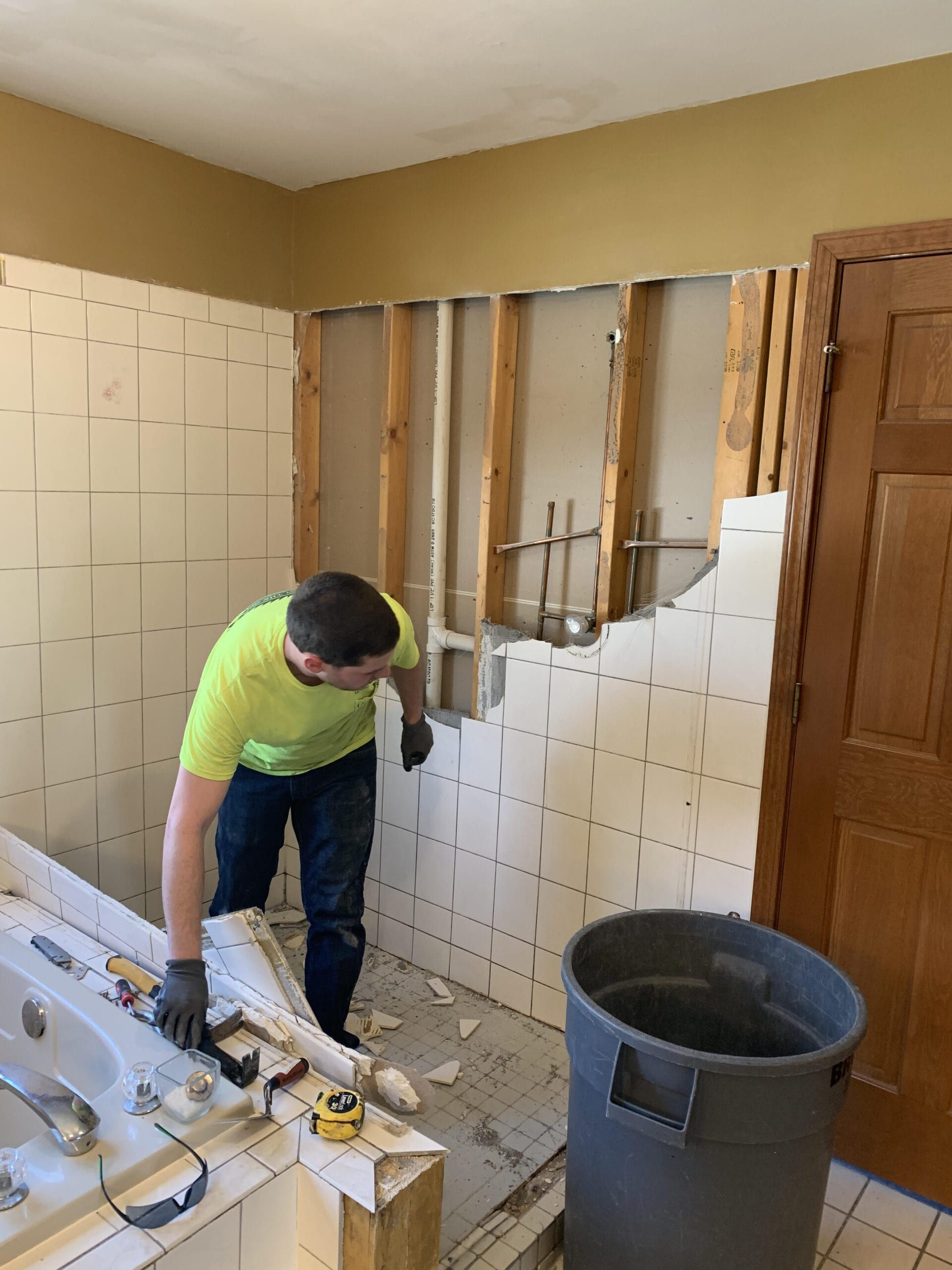 Worker in neon shirt demolishes a tiled bathroom wall beside a trash bin.