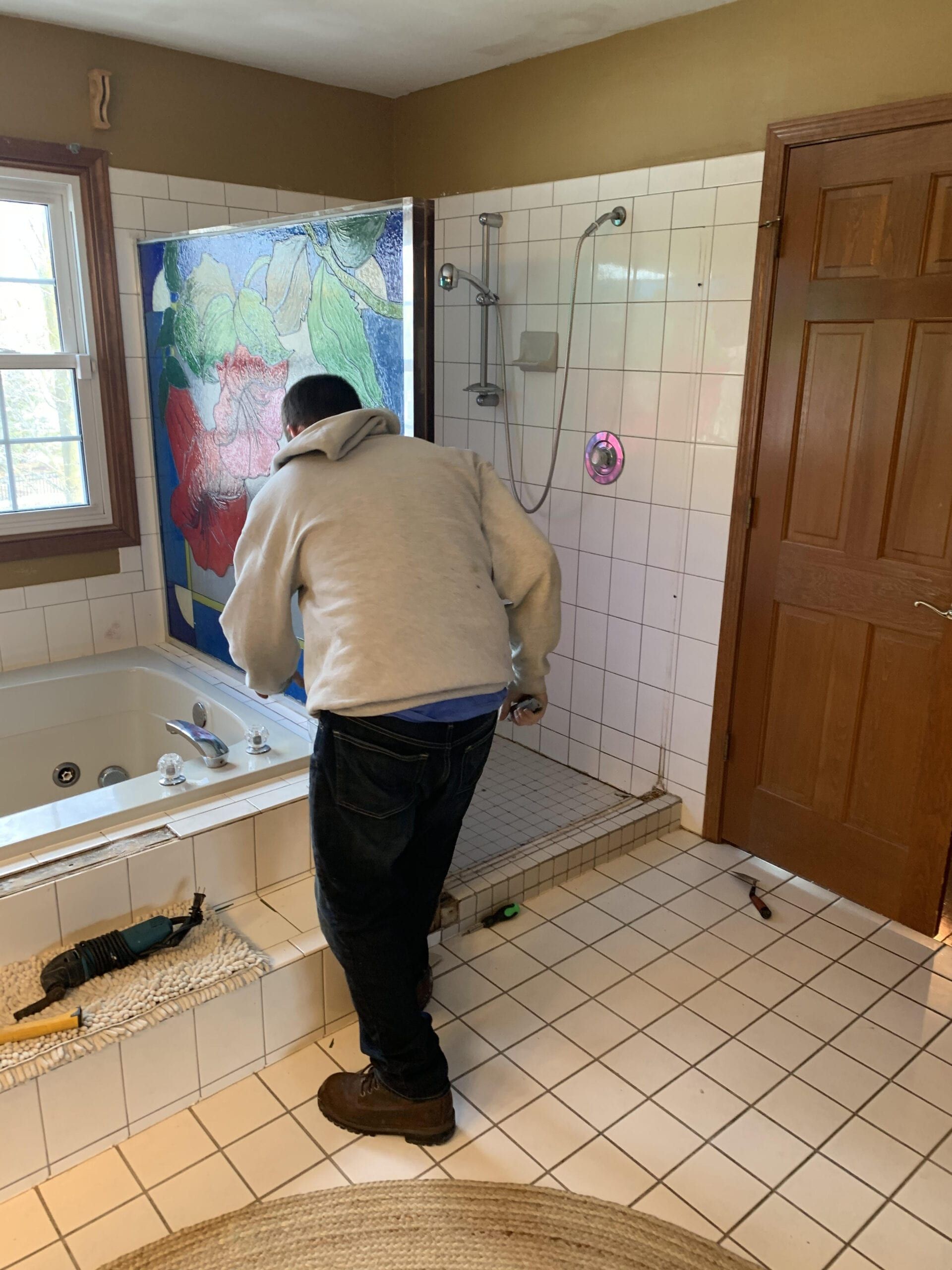 Person bending over in a tiled bathroom, cleaning near a bathtub and shower.