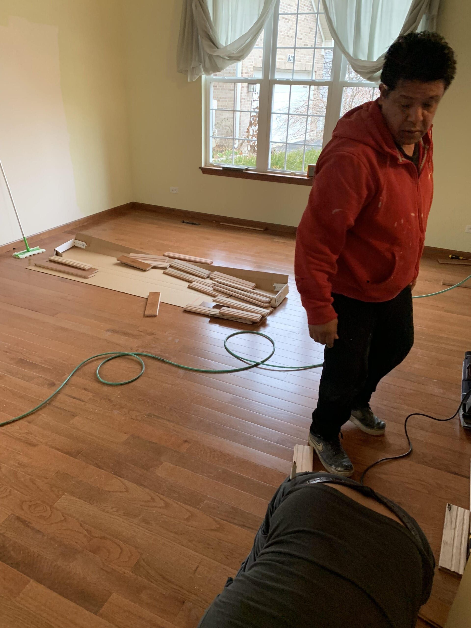Person in a red hoodie stands in a mostly empty room with wood flooring and boxes by a window.
