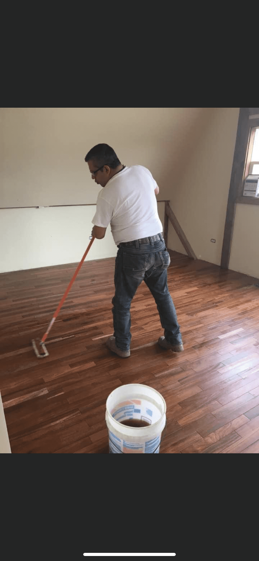 Person using a floor roller on a polished hardwood floor next to a white paint bucket.