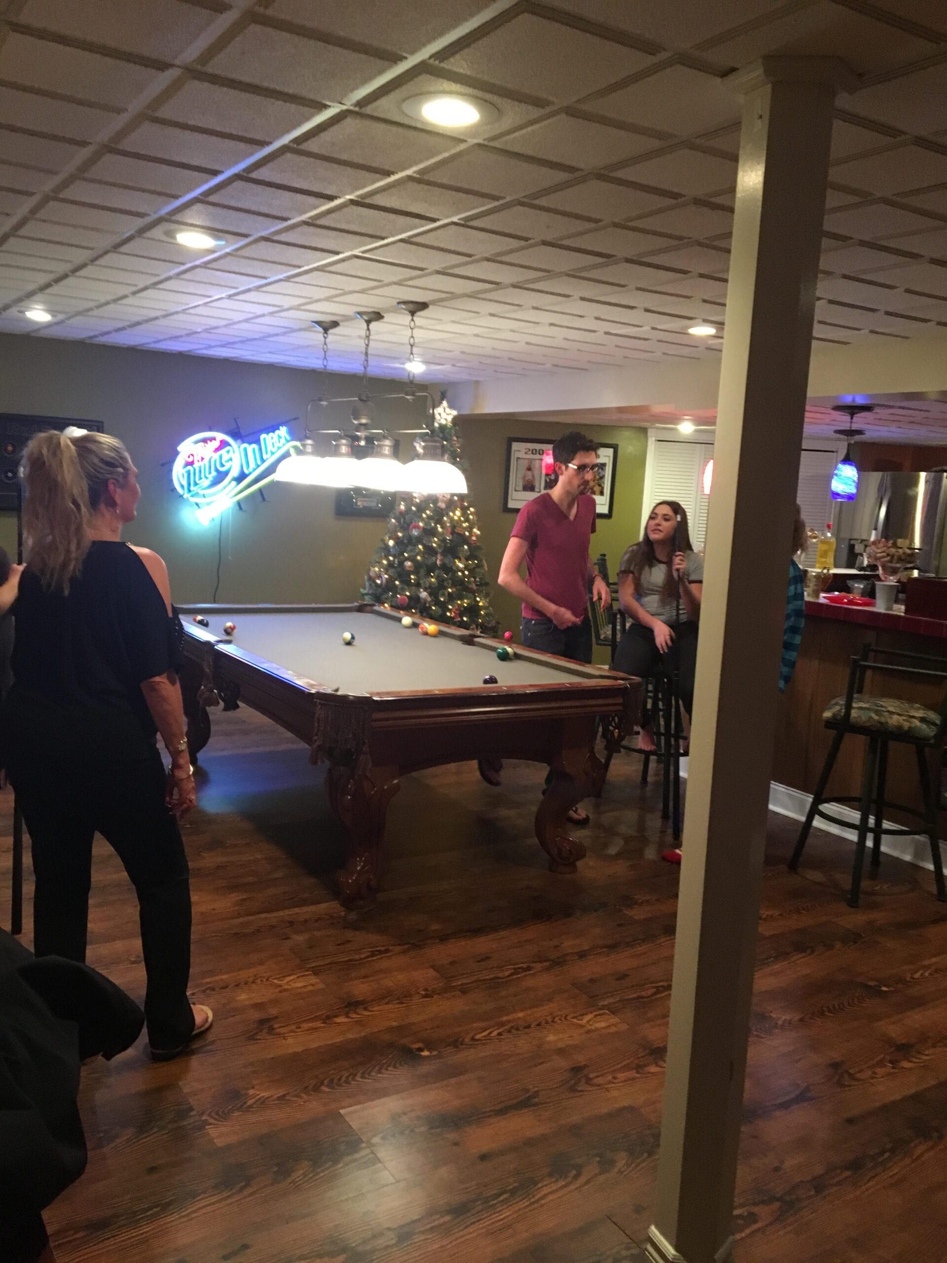 People gathered around a pool table in a dimly lit bar with a neon sign and wood floors.