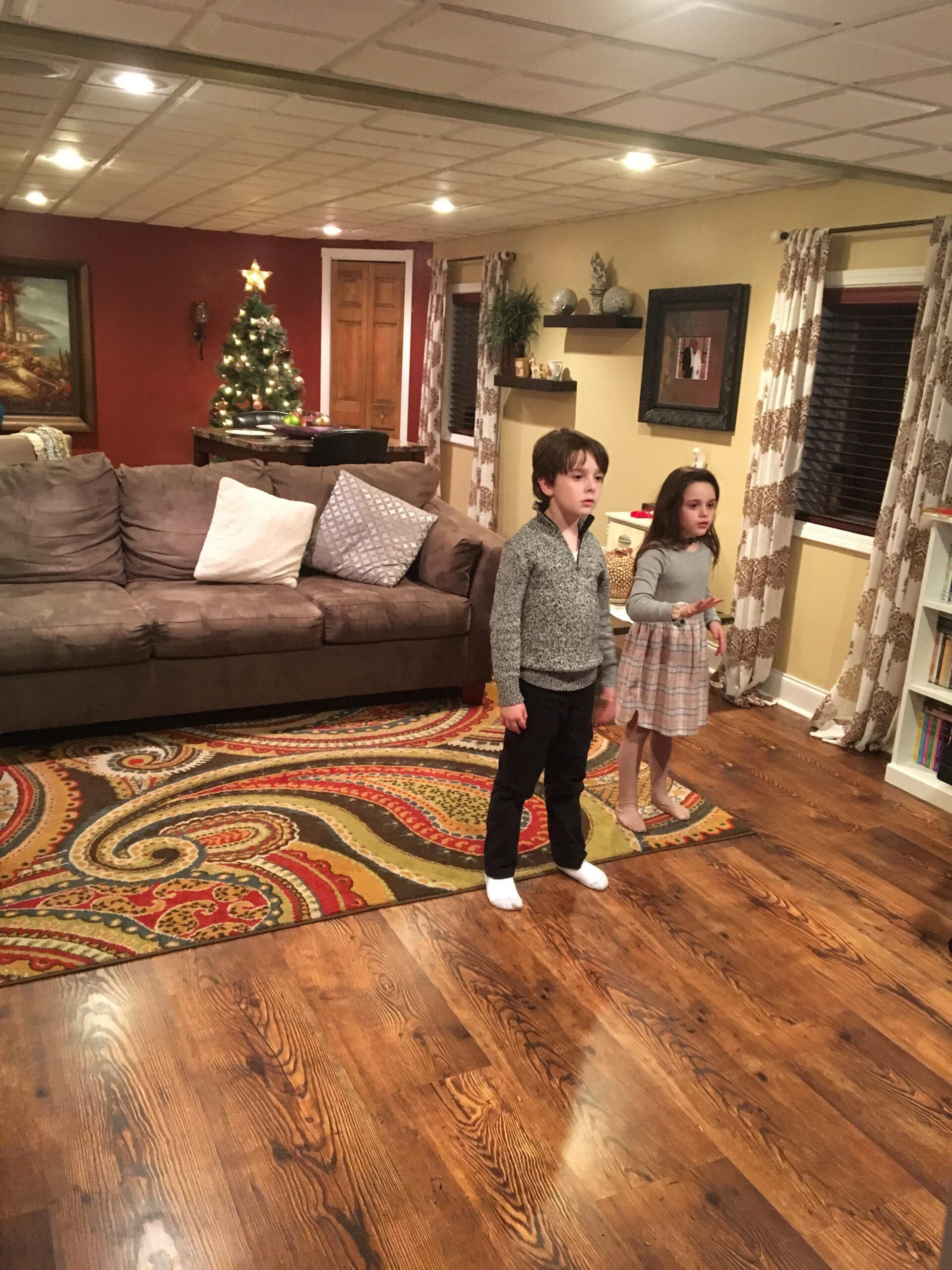 Two children standing in a cozy living room with a Christmas tree, sofa, and patterned rug.