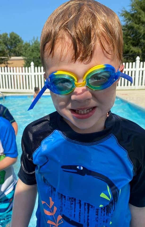 Child in blue swim shirt and goggles smiling by a pool on a sunny day