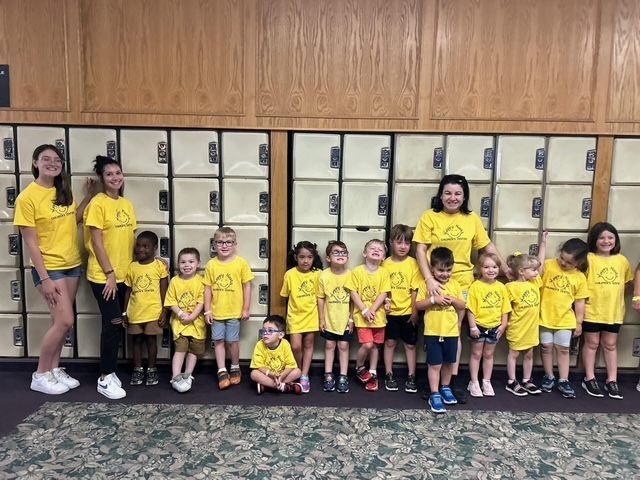 Group of children in yellow shirts posing in front of school lockers, with one adult at the left.
