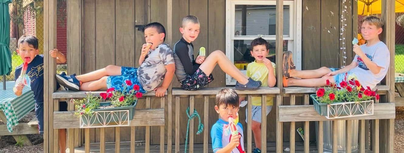 People relaxing on a porch with flower boxes, some sitting on lounge chairs and one child standing nearby