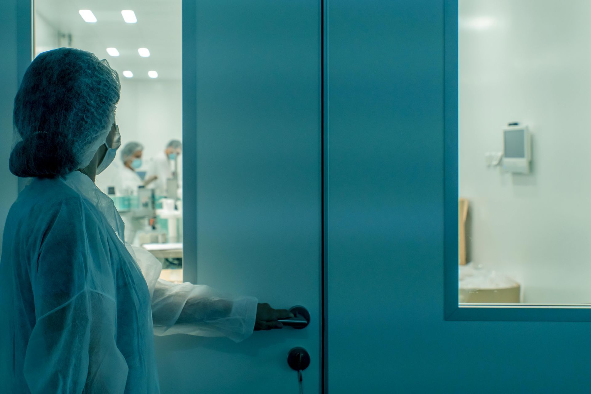 Person in protective suit opening a door to a sterile laboratory; teal lighting.