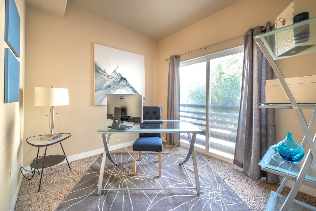 Bright in-unit home office with glass desk, monitor, blue chair, and a large window with balcony view.