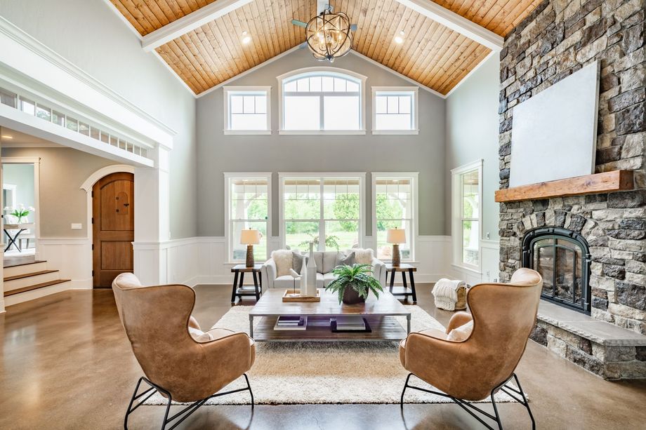 A spacious living room with high ceilings, wood paneling, and a stone fireplace. Two brown armchairs face each other n