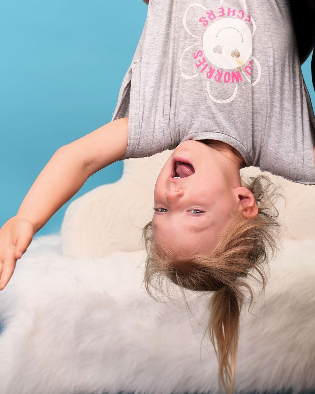 Girl upside down, laughing, wearing a grey t-shirt with a flower graphic, against a blue backdrop.