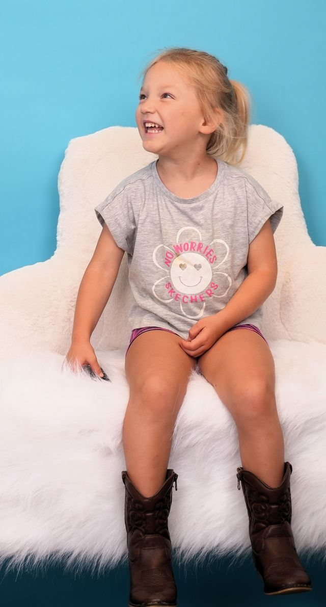 Girl in a gray shirt, sitting on fluffy white surface, wearing boots, laughing, looking upwards, blue background.