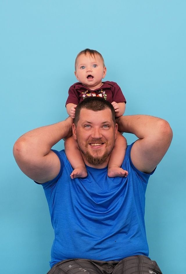 Man smiles, baby sits on his shoulders against a blue backdrop. Baby has an open mouth, wide-eyed.
