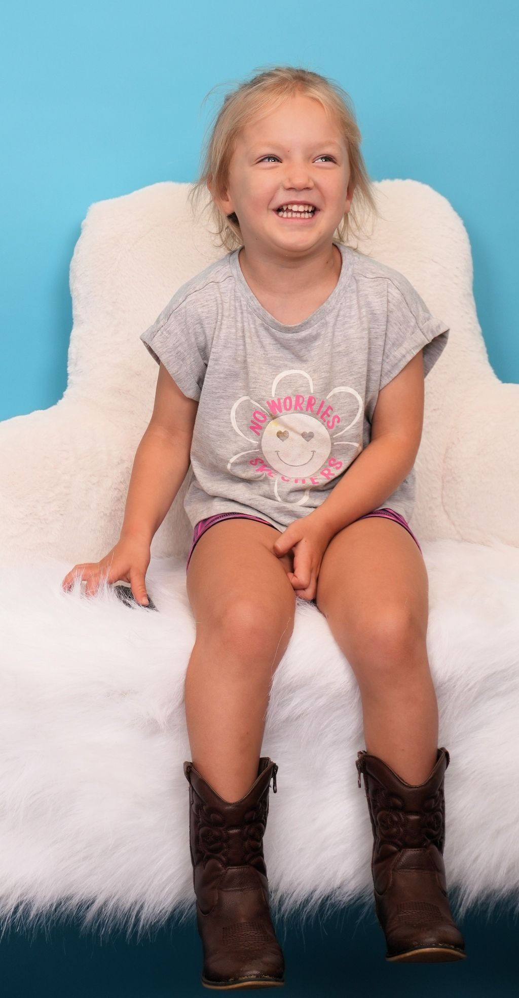 Girl sitting on a fluffy white chair, wearing a gray shirt and cowboy boots, laughing. Blue background.