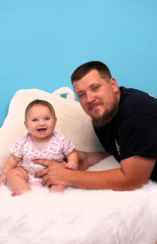 Man and baby sit on white fur, baby smiles, blue background.