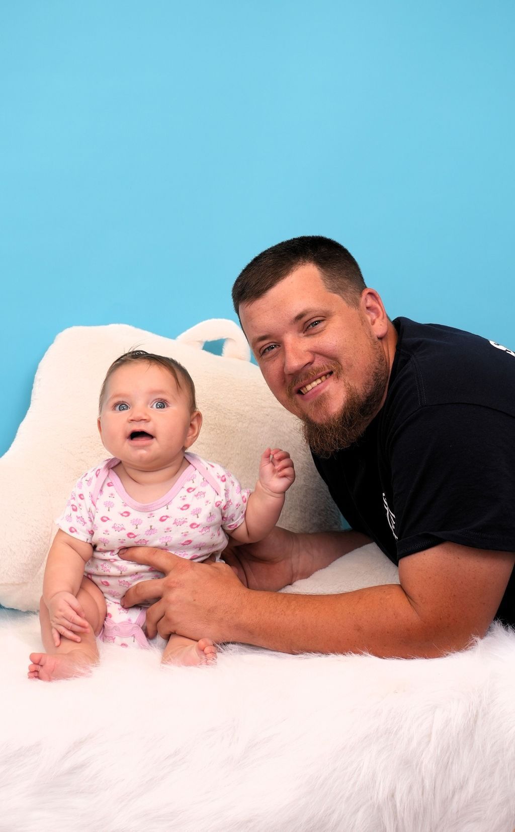 Man smiles next to baby. Baby in pink onesie, sitting on white fur against blue background.
