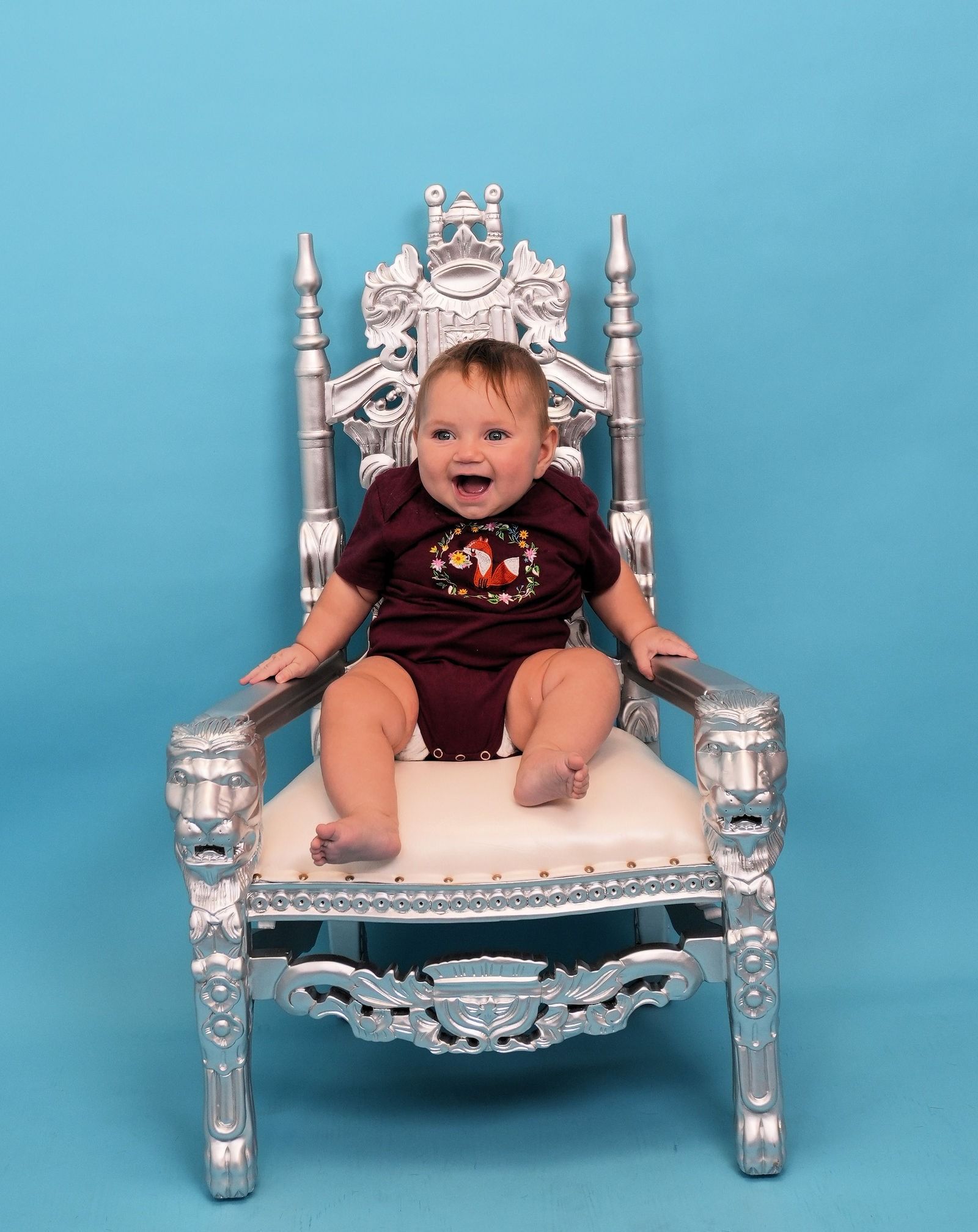 Smiling baby sits on a silver ornate throne against a blue backdrop, wearing a burgundy onesie.