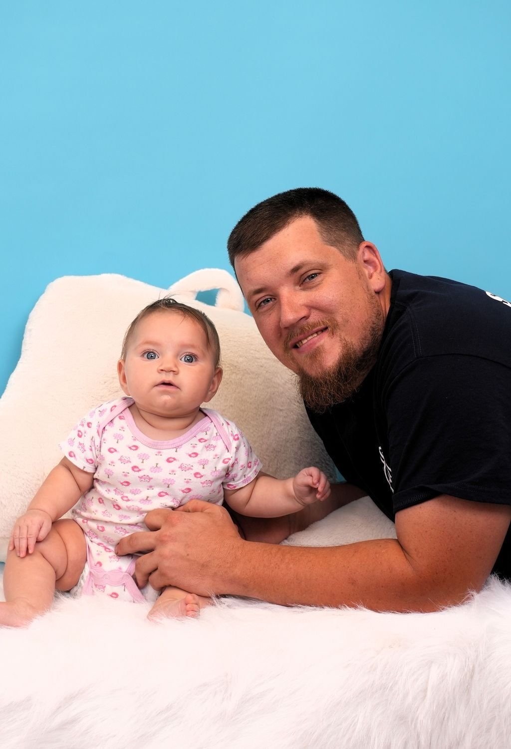 Man and baby pose on white fur, baby in pink floral onesie, blue backdrop.