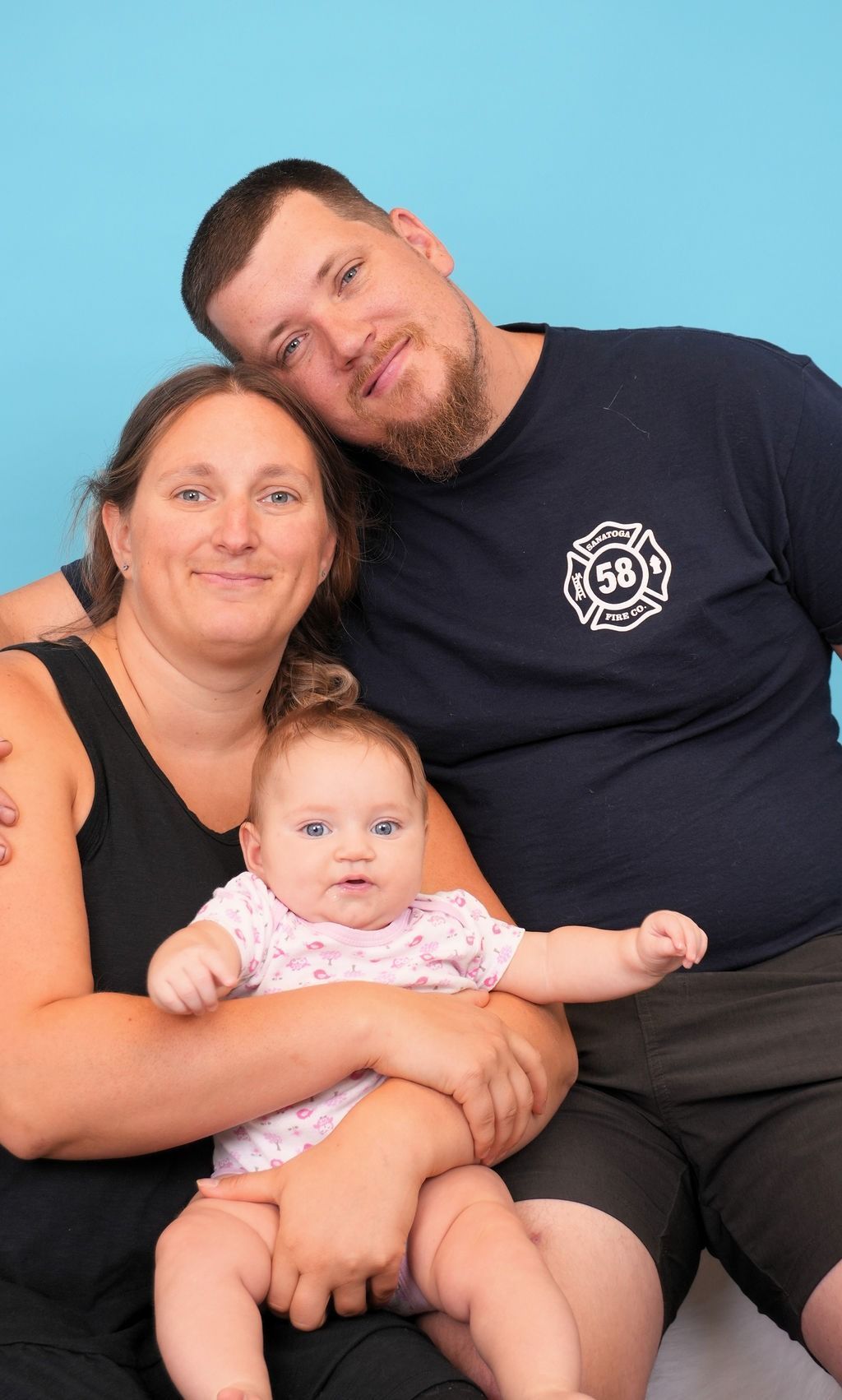 Family portrait: Smiling parents with baby; blue backdrop.