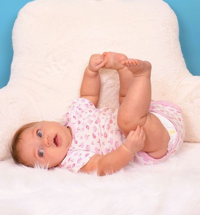 Baby in pink outfit lying on back, lifting feet and looking surprised. White fluffy backdrop.