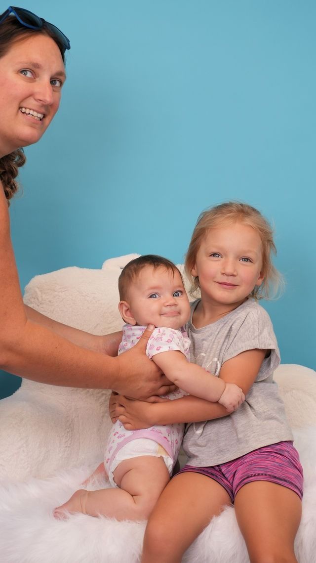 Woman holding a baby and a young girl seated on a fluffy white surface in front of a blue background.