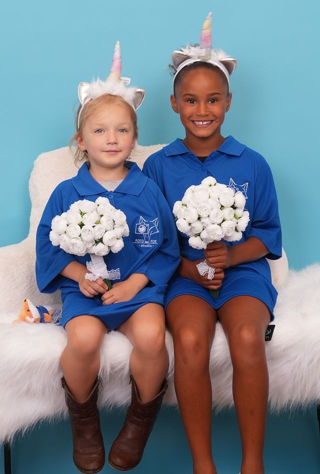 Two girls wearing unicorn headbands and holding bouquets, seated together in front of a blue backdrop.