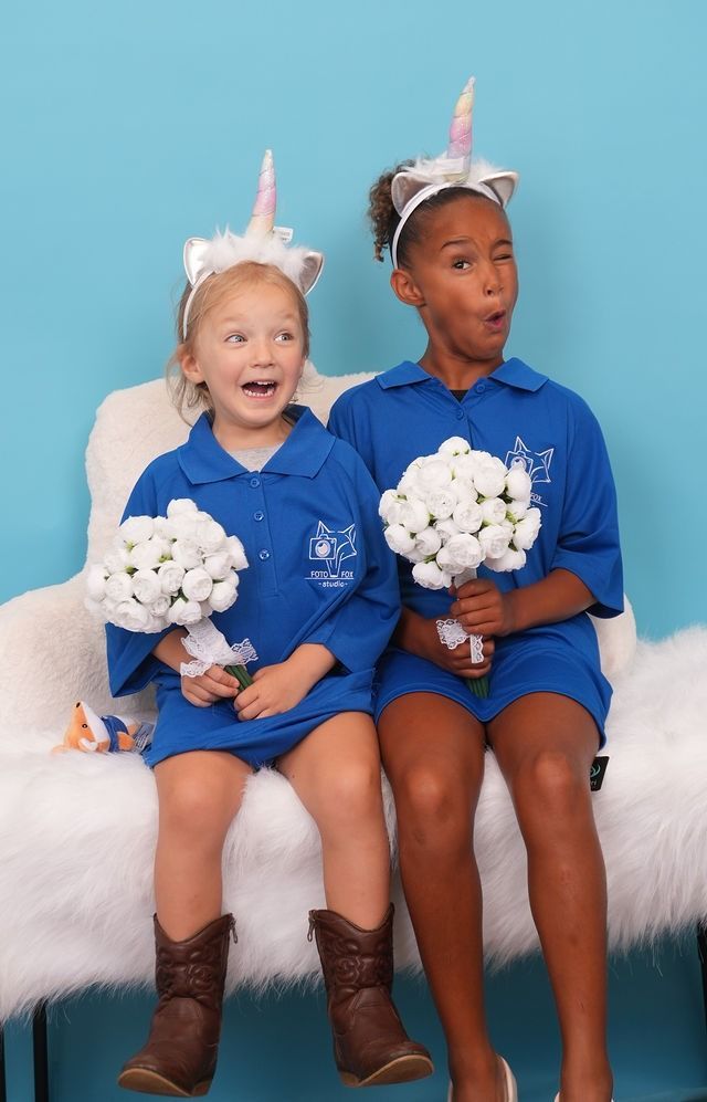 Two children in blue outfits and unicorn headbands hold bouquets, sitting on a fluffy white chair against a blue backdrop.