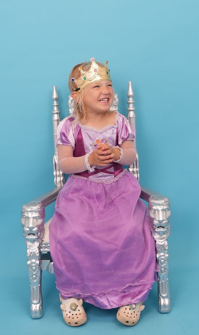 Young child in purple princess dress and gold crown sits on a silver throne, smiling.