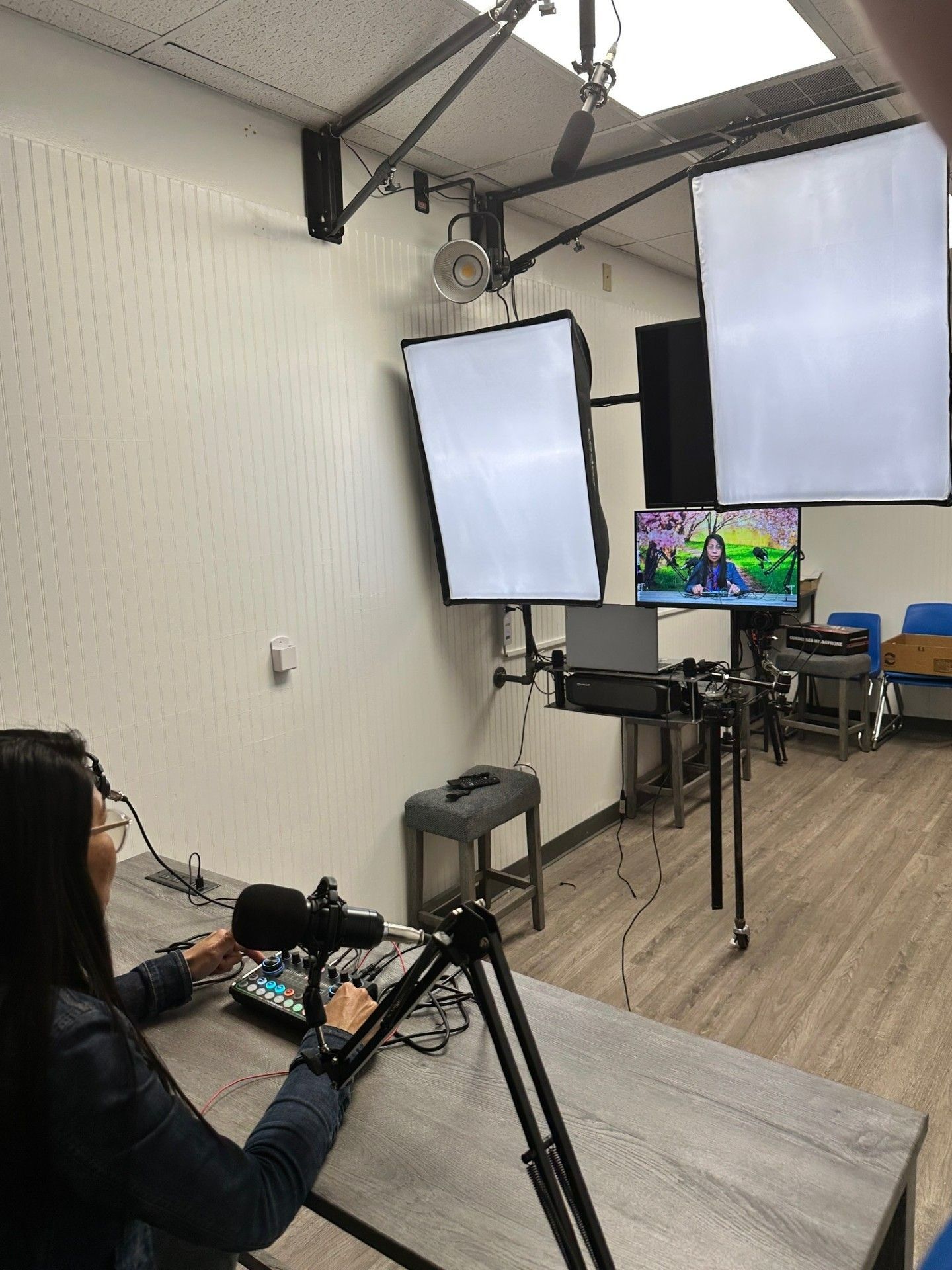 Woman at a desk with a microphone, using a soundboard in a studio. Two softboxes and a monitor are visible.