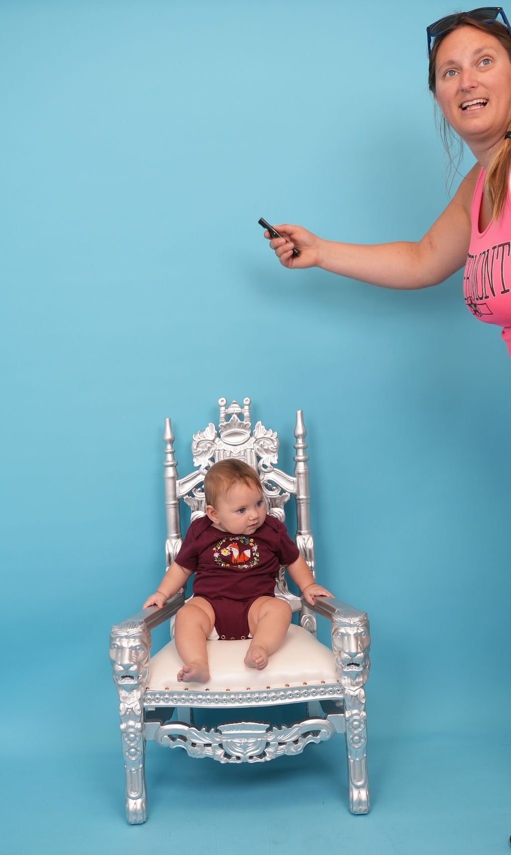 Baby in maroon onesie sits on a silver throne as woman, holding phone, leans in. Blue backdrop.