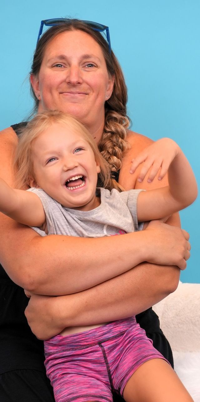 Woman hugging a laughing child. The child wears a gray shirt and pink shorts. Blue background.