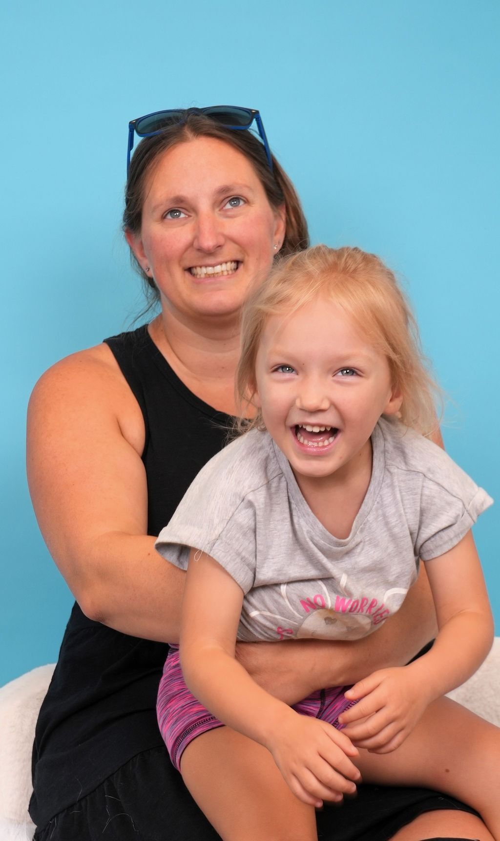 Woman holding a laughing child against a blue background. The child is wearing a gray shirt and purple shorts.