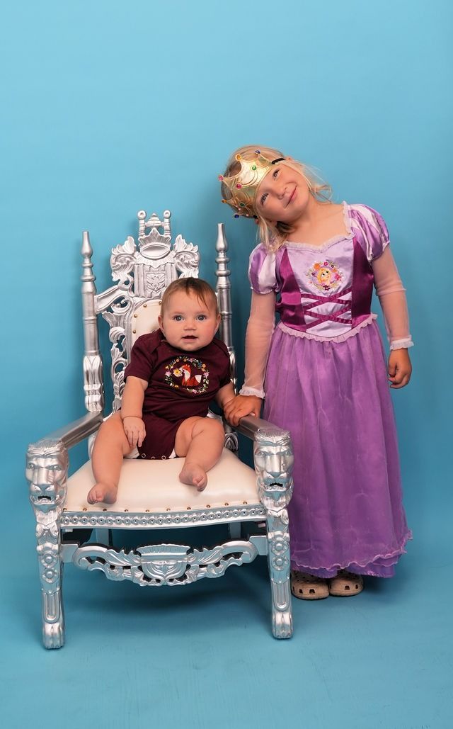 Baby sits on a silver throne; a child in princess costume stands beside it, smiling. Teal backdrop.
