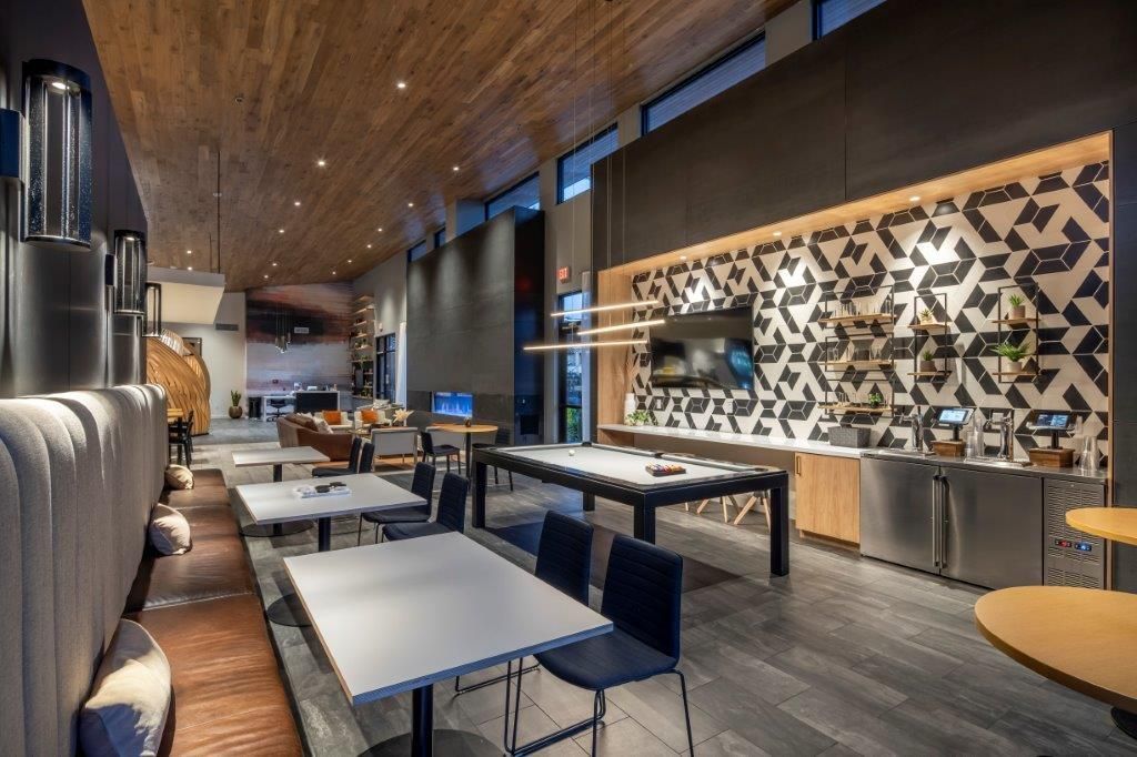 Interior of a modern apartment community lounge with tables, seating, a pool table, and a geometric wall.