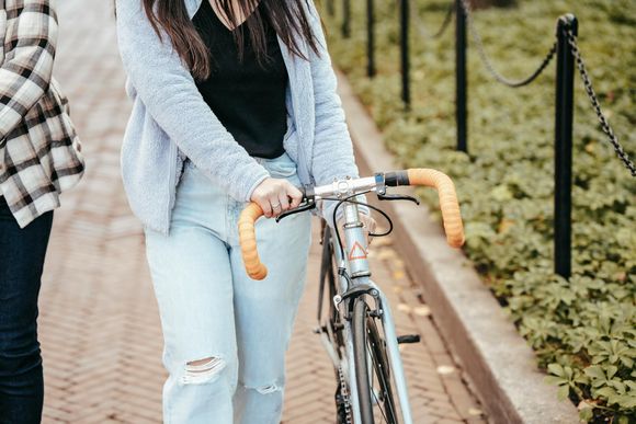 A person in a light blue jacket and light-wash jeans walking alongside a bicycle with bright orange handlebars.