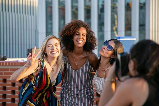 Three smiling friends pose for a photo on a rooftop terrace, with one person in the foreground taking the picture.