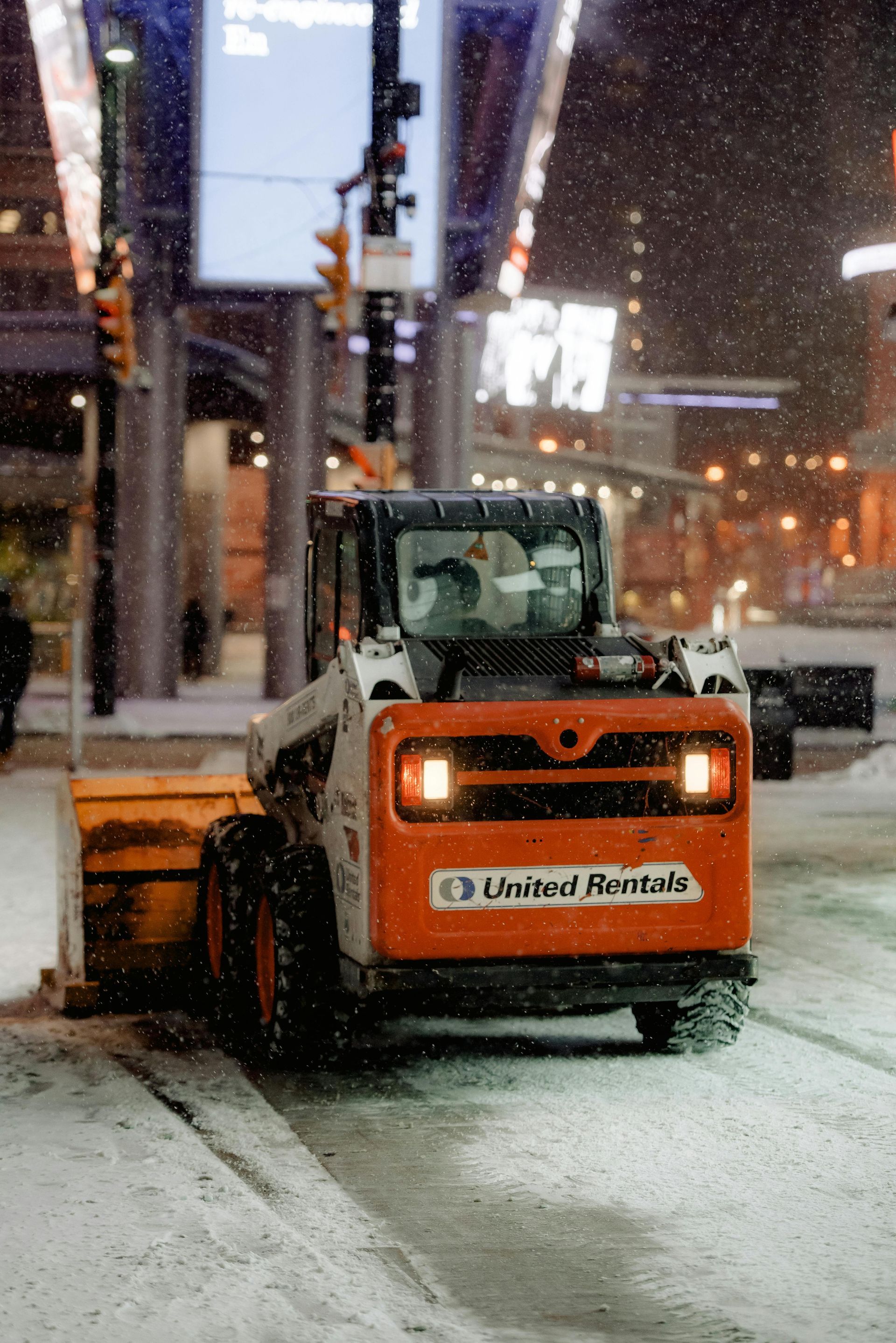 Orange United Rentals skid steer clearing snow on a city street at night