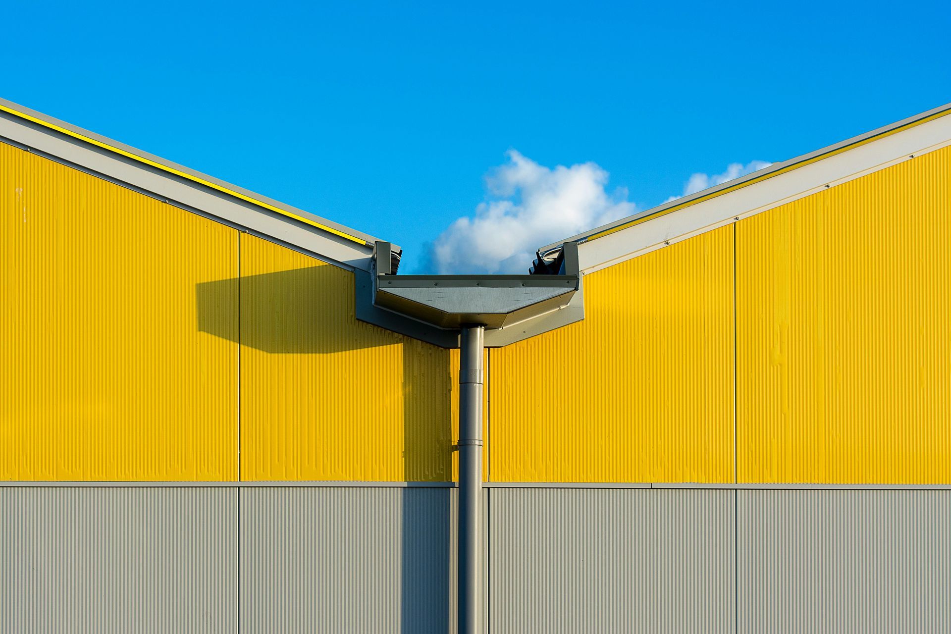 Yellow and gray industrial building roofs meeting at a central gutter under a blue sky