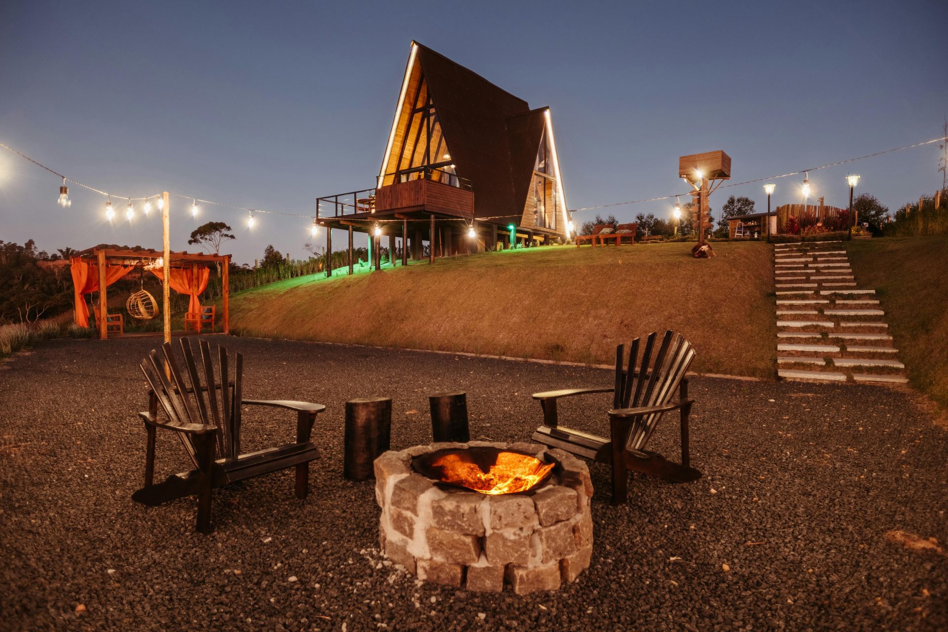 Cozy outdoor fire pit with chairs and string lights in front of a lit A-frame cabin at dusk