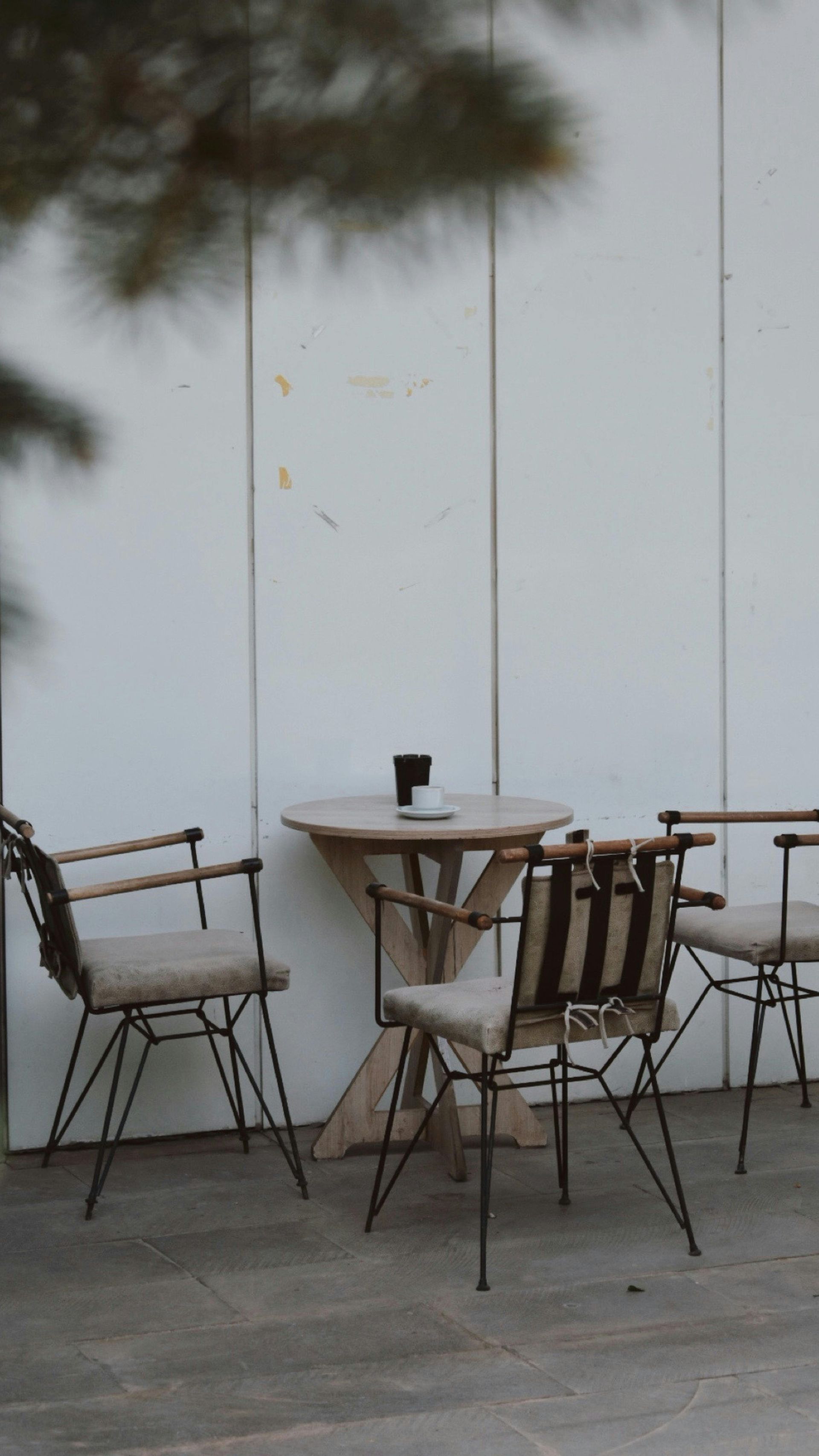Small café patio with metal chairs and a round table against a white wall, with a tree branch overhead.