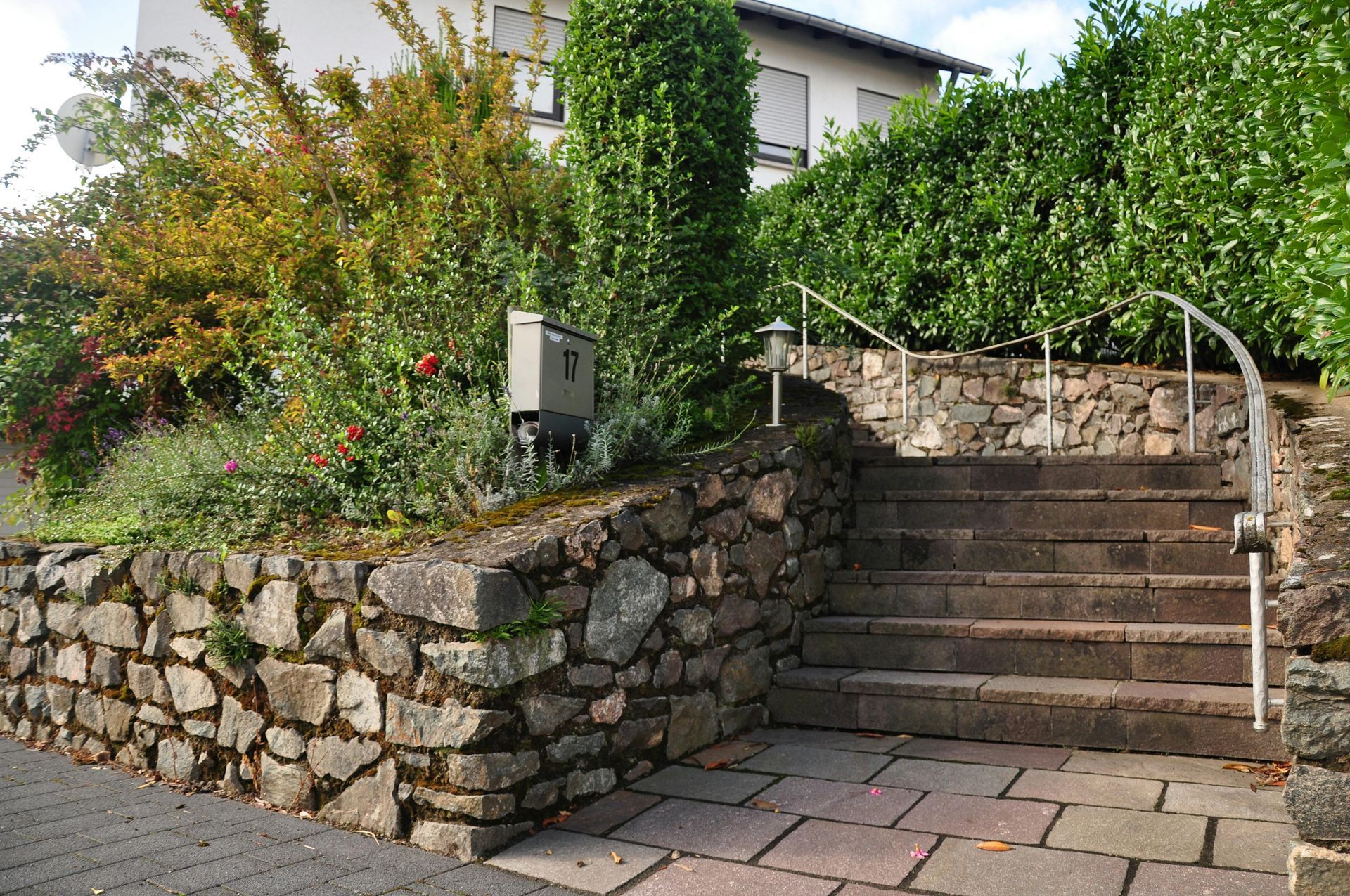 Stone steps beside a raised garden wall, with lush greenery and a house in the background.