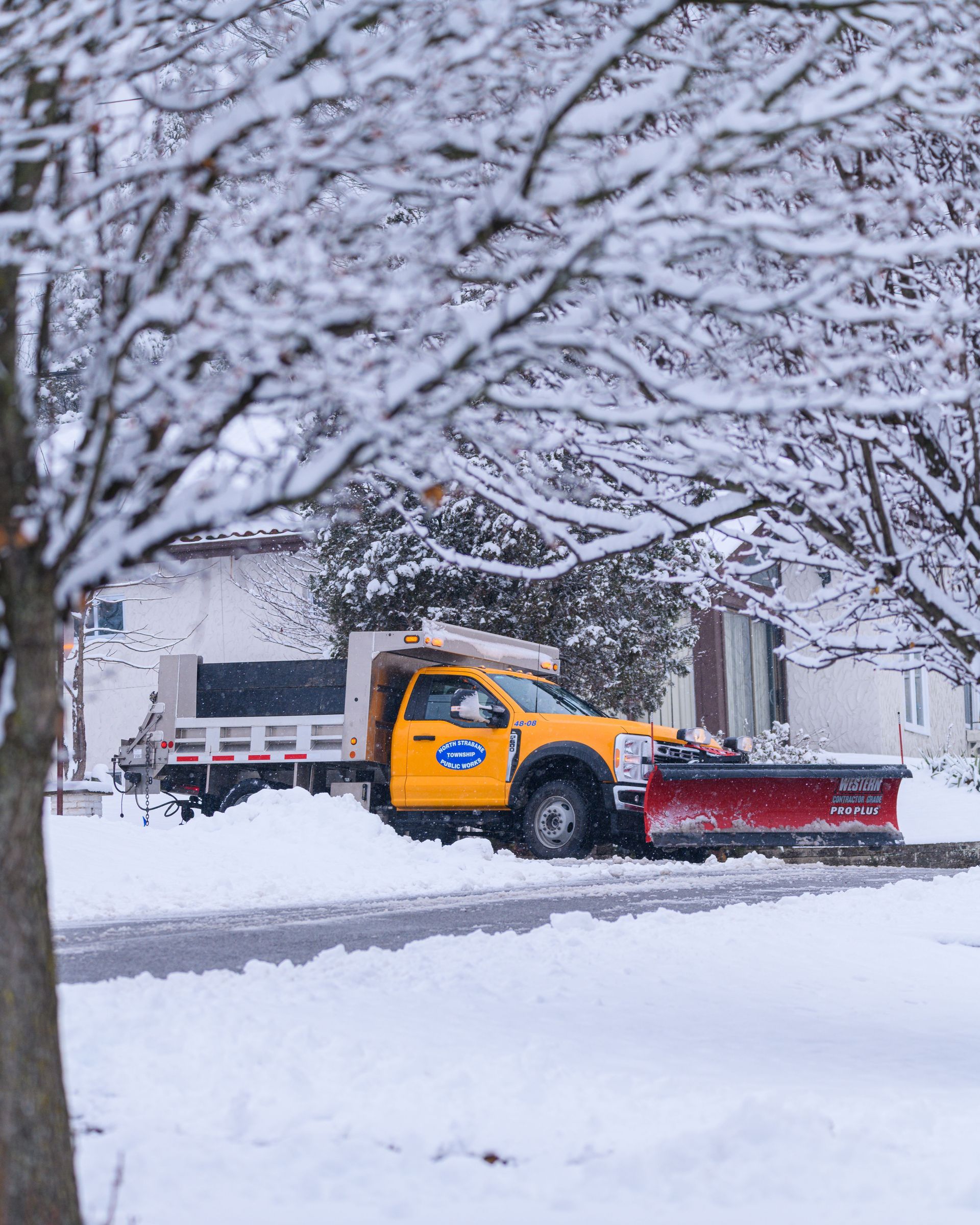 Yellow snowplow clearing a snowy residential street under leafless trees