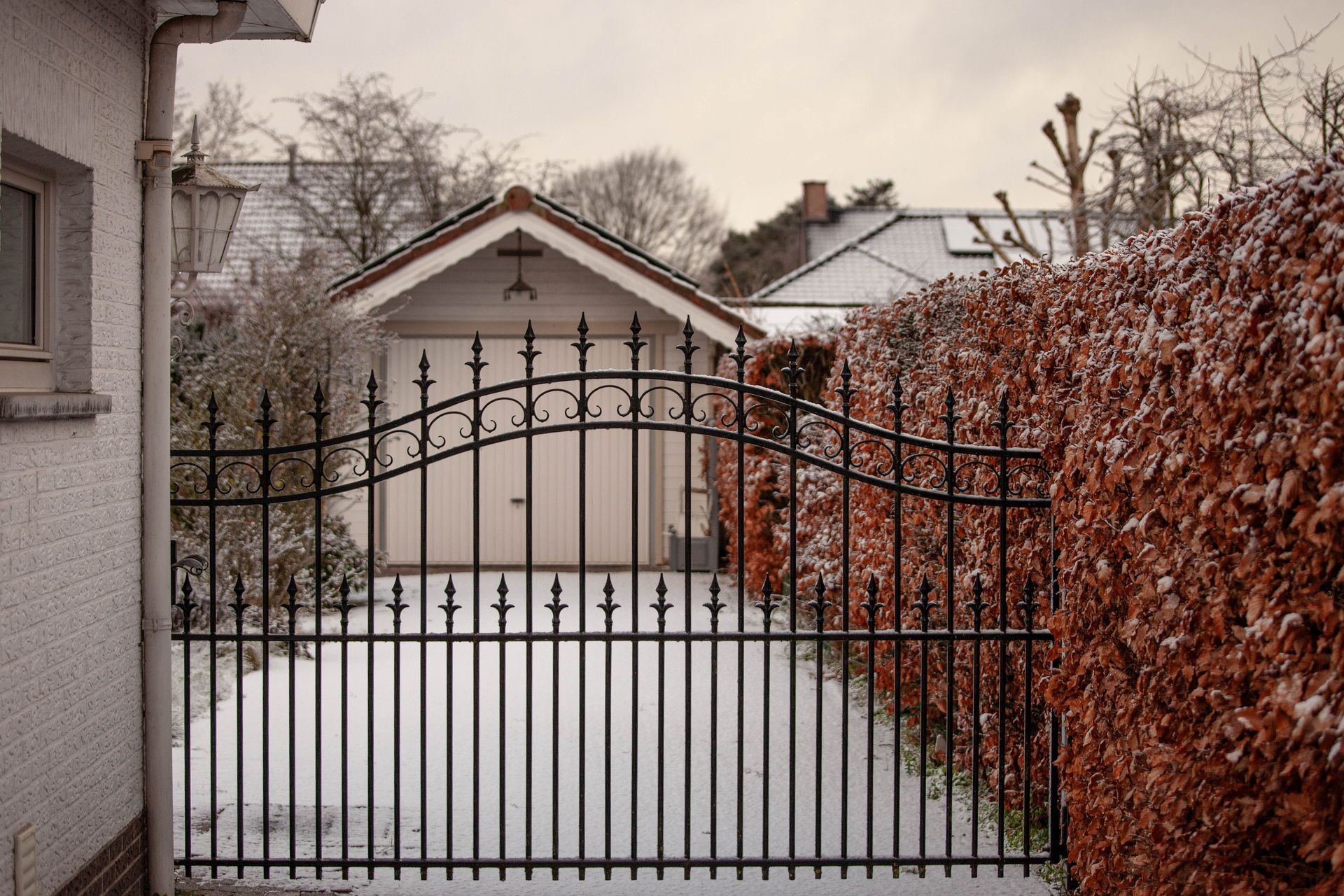 Black wrought-iron gate in front of a snowy driveway and garage, framed by a house and autumn vines