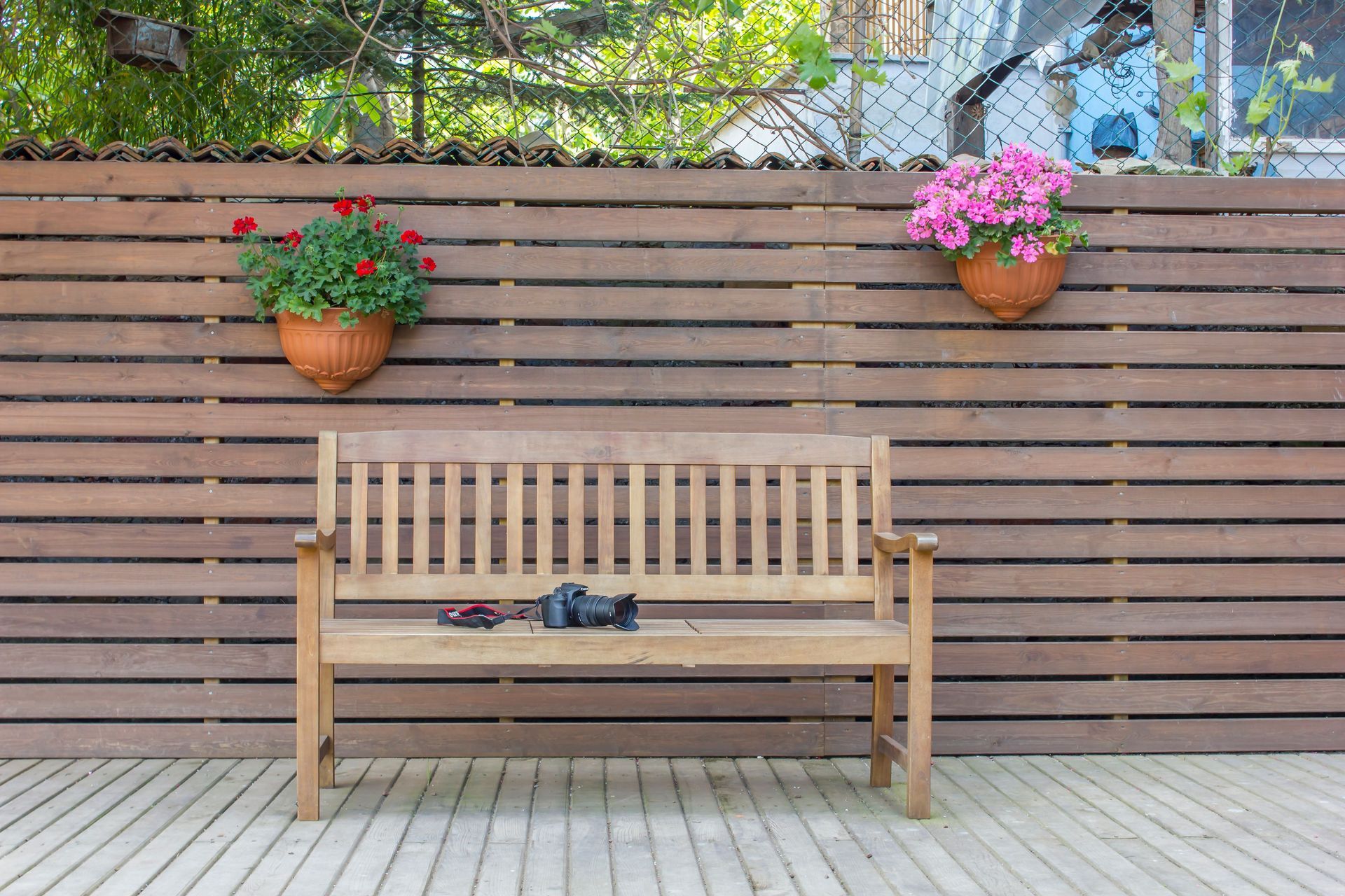 Wooden bench on a patio in front of a slatted fence with hanging flower pots and greenery above.