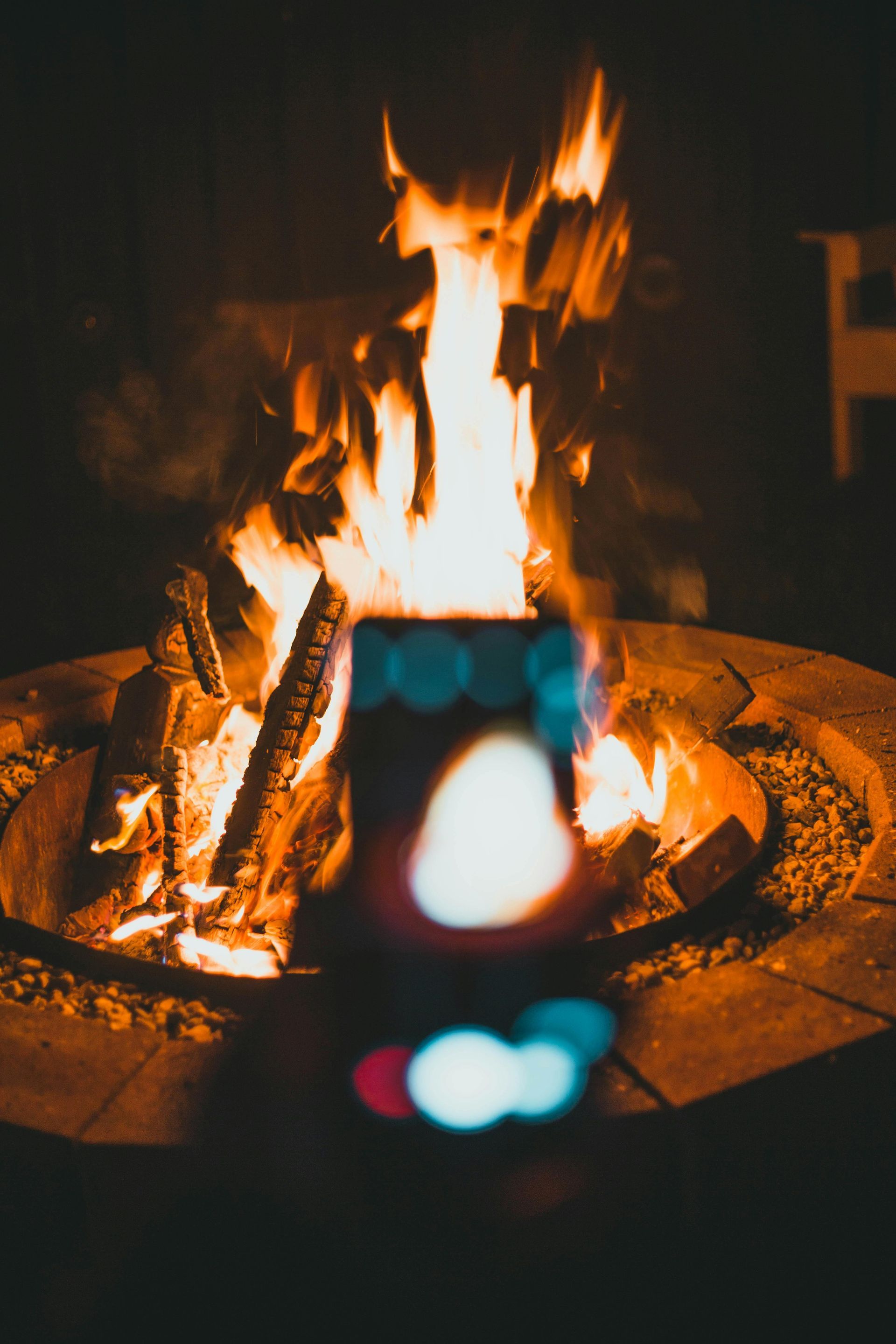 A smartphone held up to a glowing fire pit at night, with bright flames and logs in the background