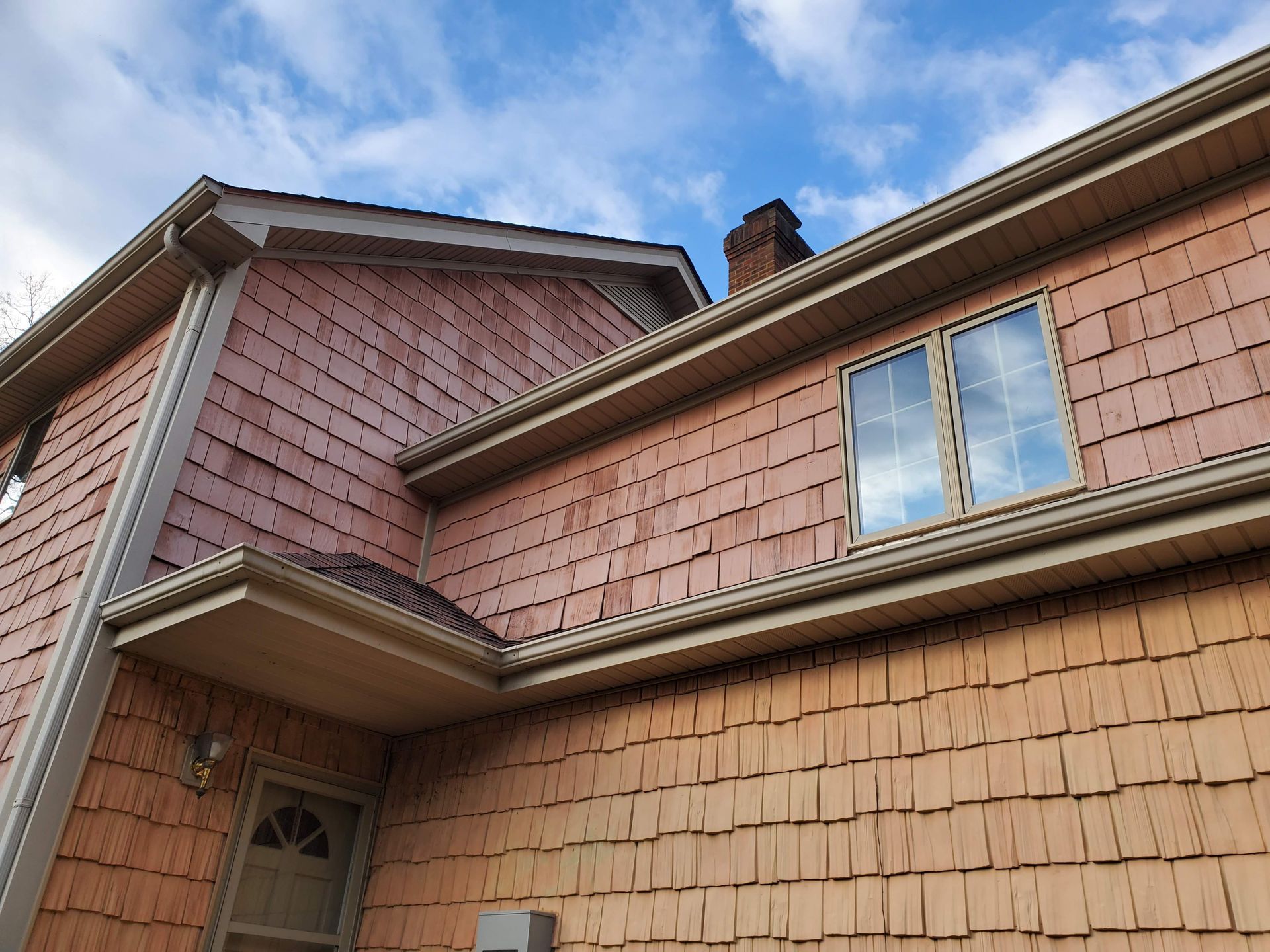 Close-up of a house roof with red shingle siding and a window against a blue sky.