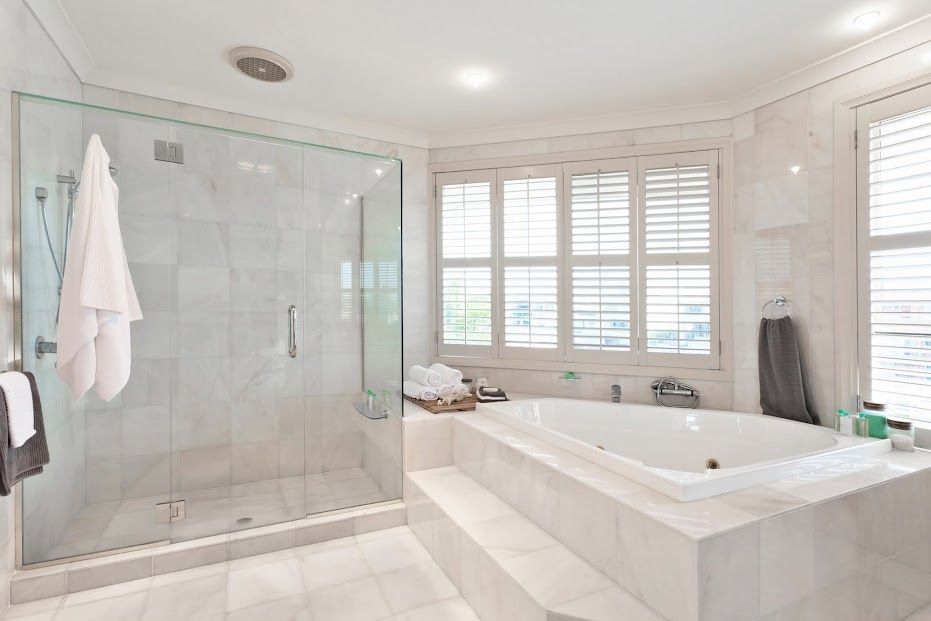 Bright white modern bathroom with glass shower, soaking tub, and window shutters