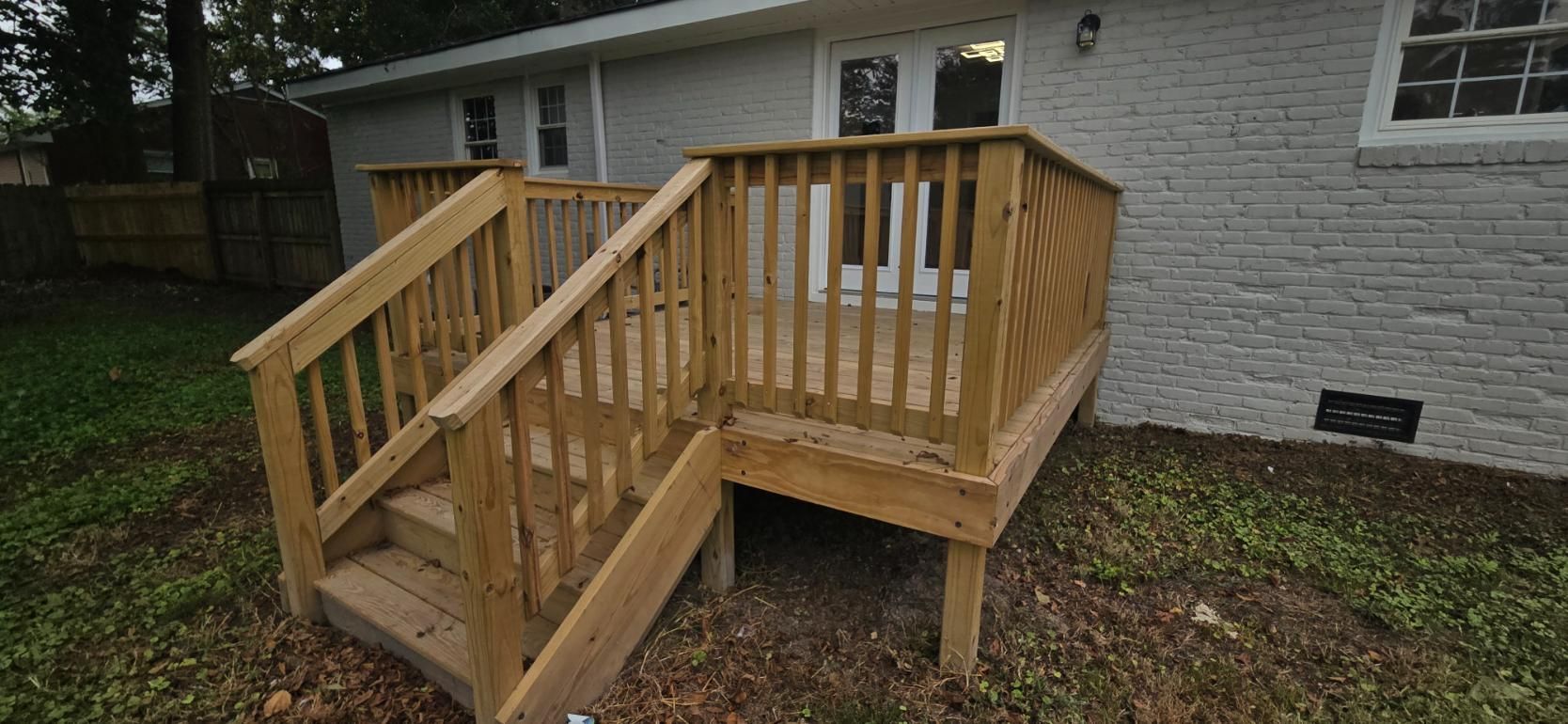 Wooden deck with stairs attached to a light gray house with a brick wall.