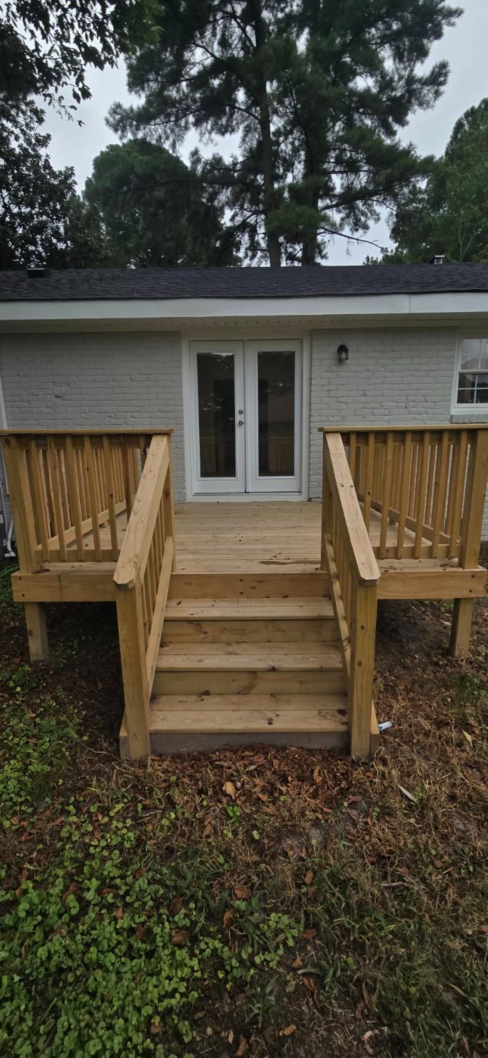 Wooden deck with stairs leading to a building with double doors, set in a yard with trees and overcast sky.
