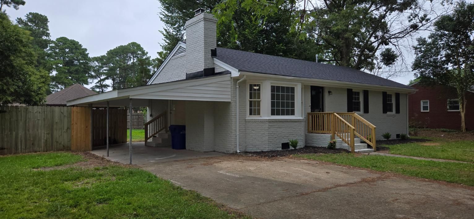 A gray-painted house with a black roof and a carport, trees in the background.