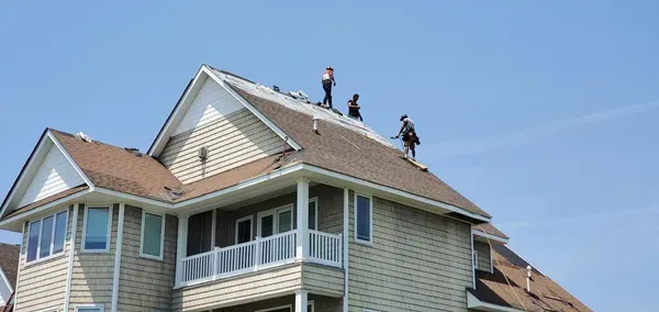 Three people on a rooftop repair a house on a clear, sunny day.